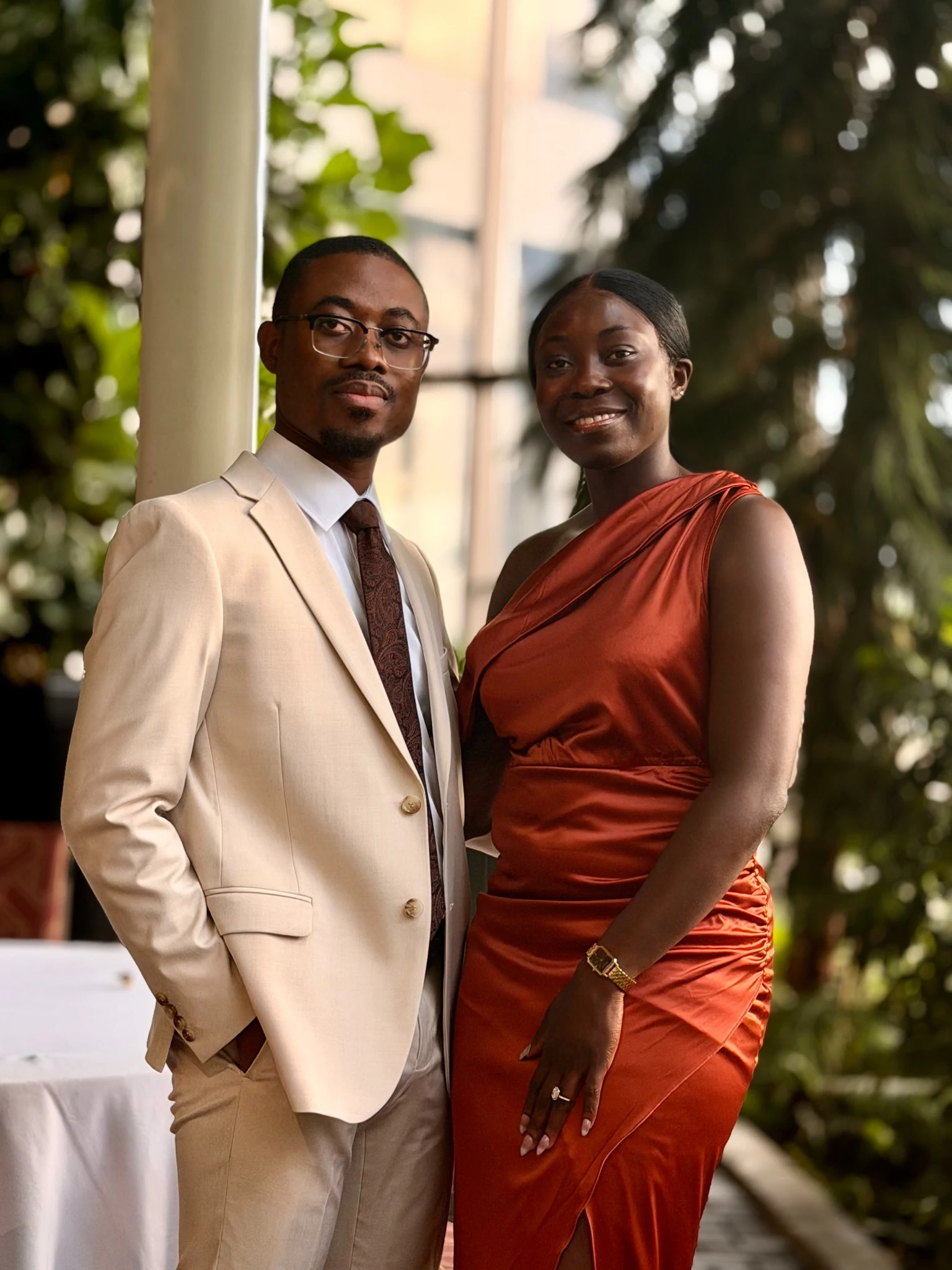 Frederick in a cream suit beside Veronica in a rust satin dress, standing together outdoors